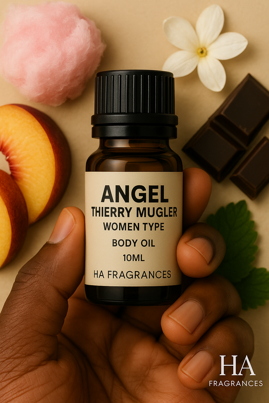 Dark-skinned hand holding a 10ml HA Fragrances Angel Thierry Mugler Women Type body oil bottle, surrounded by chocolate pieces, cotton candy, peach slices, jasmine flower, and green leaf on a beige background.