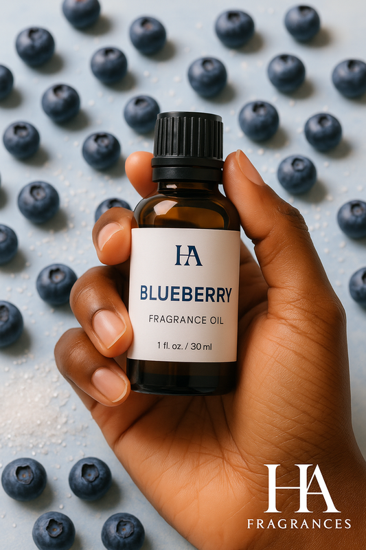 A dark-skinned hand holding a bottle of HA Fragrances Blueberry Fragrance Oil, with fresh blueberries and sugar crystals in the background.