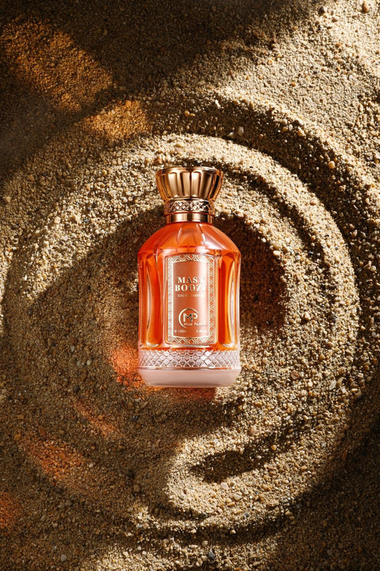 Mast Boozy Eau de Parfum by Bharara – luxurious amber bottle with gold cap resting on sand, representing a warm vanilla-caramel boozy fragrance.