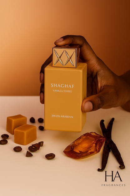 Dark-skinned hand holding Swiss Arabian Shaghaf Vanilla Toffee Eau De Parfum bottle, surrounded by toffee pieces, coffee beans, vanilla pods, and warm amber tones with HA Fragrances logo.