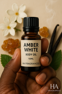 Dark-skinned hand holding a 10ml bottle of Amber White Body Oil by HA Fragrances, surrounded by vanilla beans, jasmine flowers, amber resin, and mint leaves on a beige background.
